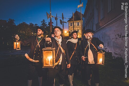 Eine Gruppe von Nachtwächtern steht bei Nacht mit ihren Leuchten und Hellebarden in der Altstadt Bambergs